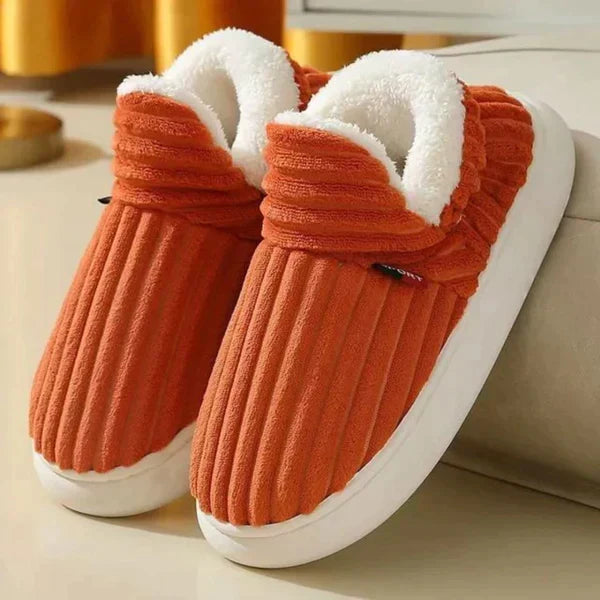Oranje geribbelde pluchen pantoffels met witte fleece voering en dikke witte zool, schuin neergezet naast een bank. 6