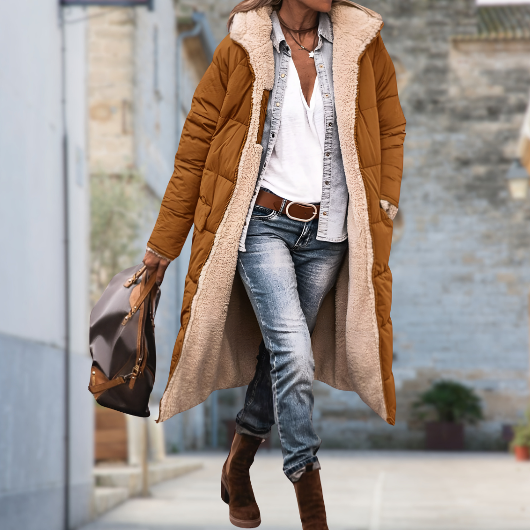 Model loopt door een straat in de lange gewatteerde jas in bruin-oranje, open gedragen met beige fleecevoering aan de binnenkant. 9