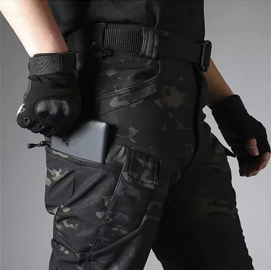 Detail van de camouflagebroek met rits-zakken op het been; een telefoon zit half in het vak, de riem is zichtbaar. 19