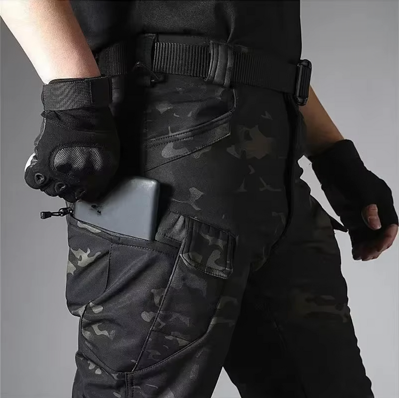 Detail van de camouflagebroek met rits-zakken op het been; een telefoon zit half in het vak, de riem is zichtbaar. 19